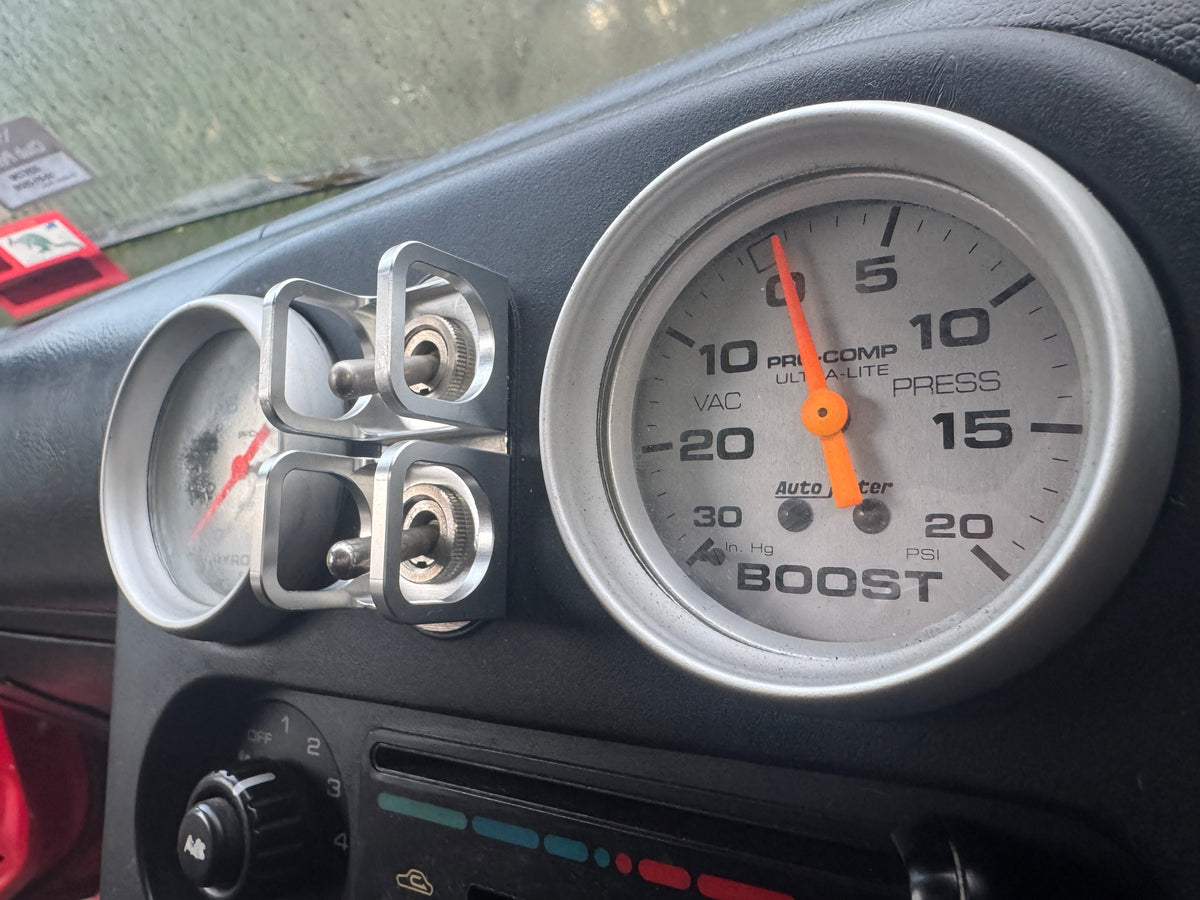 Duskie silver billet aluminum toggle switch guard with two switches, mounted next to a boost gauge in a vehicle dashboard.